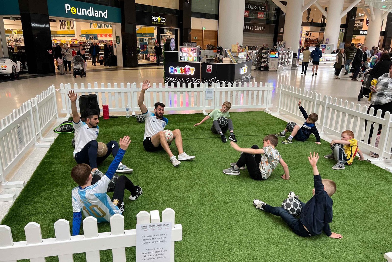 Football freestyle coach leading seated tricks workshop with kids in indoor shopping centre setup